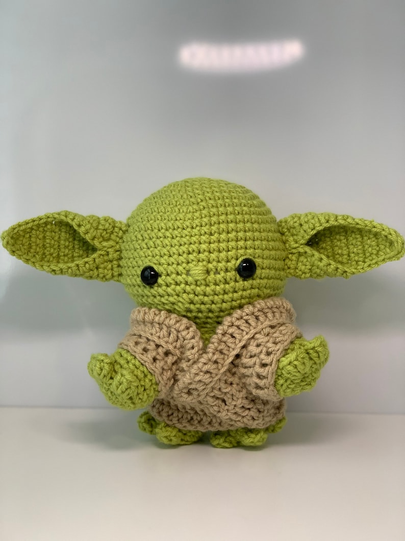 May include: A green crocheted plush toy with large ears and a brown crocheted vest. The toy has black eyes and a small mouth.