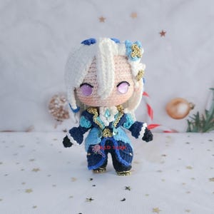 Puede incluir: Una muñeca de ganchillo hecha a mano con cabello blanco con detalles azules, piel clara y ojos morados. La figura viste un atuendo detallado en azul marino y azul claro con adornos dorados y pequeñas flores azules. El texto RED YARN es visible.