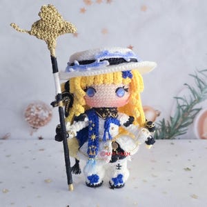 Puede incluir: Una muñeca de ganchillo hecha a mano con largo cabello amarillo, que lleva un sombrero blanco con detalles azules y un conjunto blanco con detalles en azul y negro. La muñeca sostiene un bastón alto con un elemento dorado y muestra el texto RED YARN.