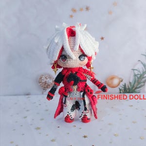 May include: A handcrafted crochet doll featuring white hair with a red streak, wearing a red, black, and white outfit with gold star-shaped earrings. The doll has large dark eyes and the text FINISHED DOLL is printed in red on the right side.