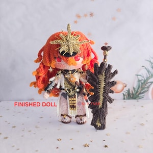 May include: A handmade crochet doll with orange hair and a gold crown, holding a brown and gold crochet weapon. The doll is wearing a white and gold outfit. The text "FINISHED DOLL" is visible on the left side of the image.