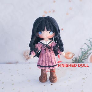 May include: A handmade crochet doll with black hair and blue eyes, wearing a pink dress with black trim and brown boots. The doll has a white collar and a small bow. The text "FINISHED DOLL" is visible.