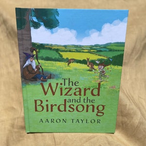 May include: A hardcover book titled "The Wizard and the Birdsong" by Aaron Taylor. The cover features an illustration of a wizard, a child, and kangaroos in a green field under a blue sky.