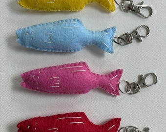 May include: Four felt fish keychains in yellow, blue, pink, and red. Each fish has silver stitching details and a metal clasp. The keychains are arranged vertically, showcasing the variety of colors and designs.