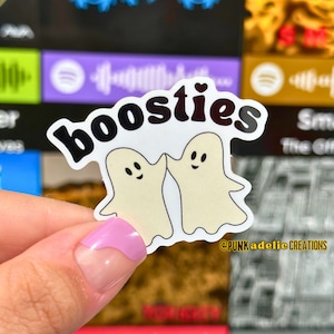 May include: A white sticker with two cartoon ghosts high-fiving each other. The sticker says "boo-sties" in black letters with a small heart above the "s". The sticker is held by a hand with pink nail polish.