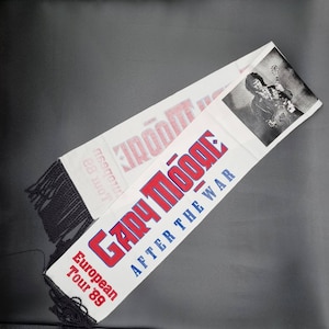 以下が含まれることがあります： 「Gary Moore - After the War」と「European Tour '89」の文字が書かれた白と赤の スカーフ。右上の隅には、ギターを弾くゲイリー・ムーアの白黒写真が写っている。