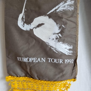 Puede incluir: Una bufanda de tela marr&oacute;n con un dise&ntilde;o de p&aacute;jaro blanco y el texto "EUROPEAN TOUR 1992". La bufanda tiene una franja de crochet amarilla.