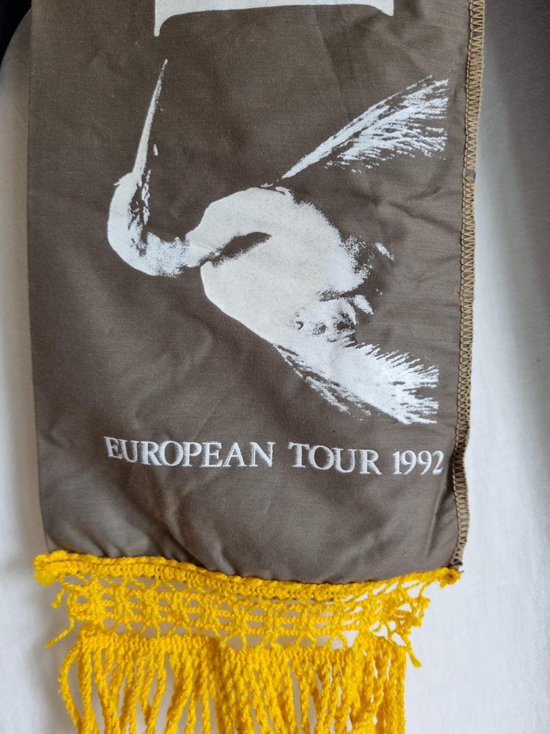 Puede incluir: Una bufanda de tela marr&oacute;n con un dise&ntilde;o de p&aacute;jaro blanco y el texto "EUROPEAN TOUR 1992". La bufanda tiene una franja de crochet amarilla.
