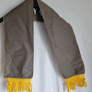 Puede incluir: Una bufanda de tela gris con flecos amarillos en los extremos. La bufanda est&aacute; colgada de una percha.