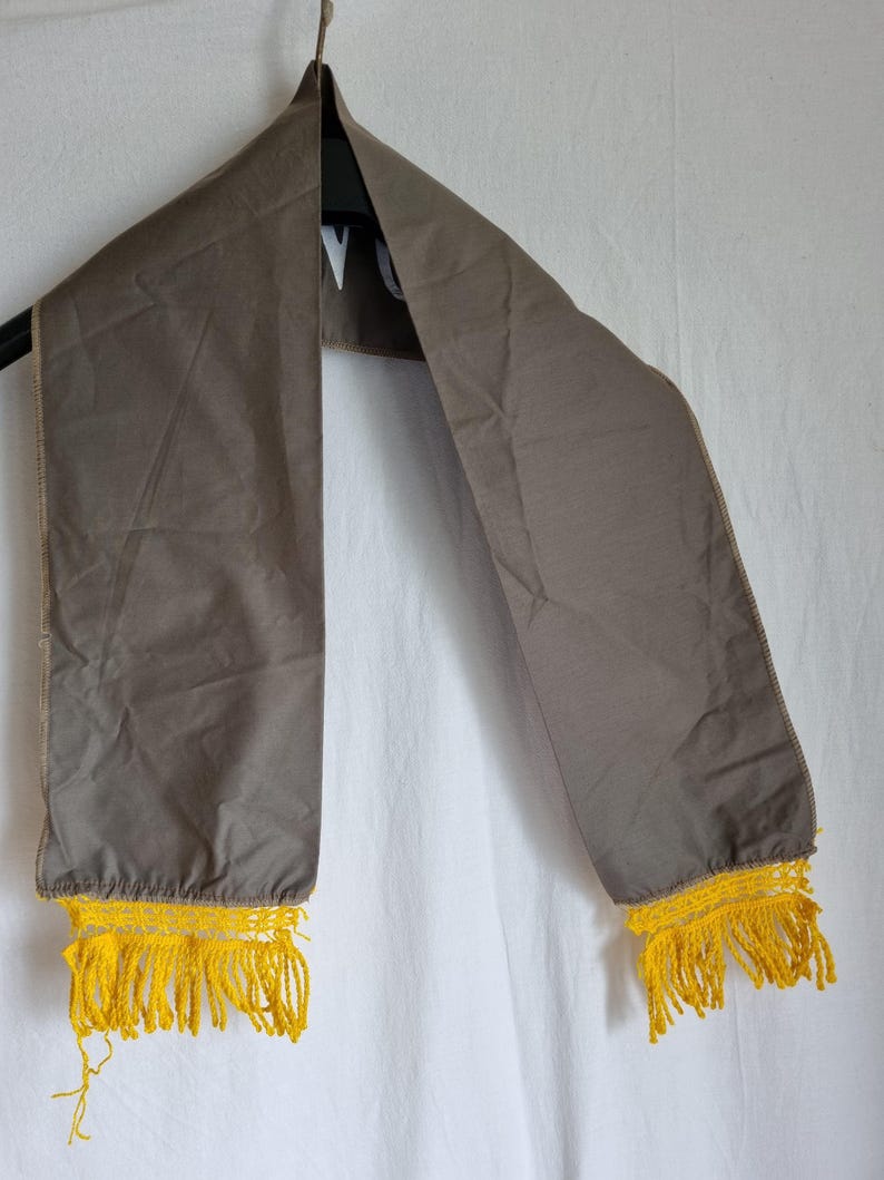 Puede incluir: Una bufanda de tela gris con flecos amarillos en los extremos. La bufanda est&aacute; colgada de una percha.