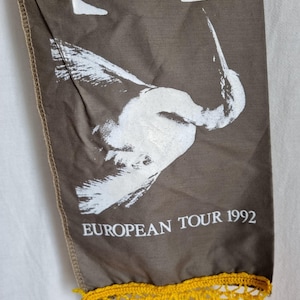 Puede incluir: Una pancarta de tela marr&oacute;n con un dise&ntilde;o de p&aacute;jaro blanco y el texto "EUROPEAN TOUR 1992". La pancarta tiene una franja de crochet amarilla.