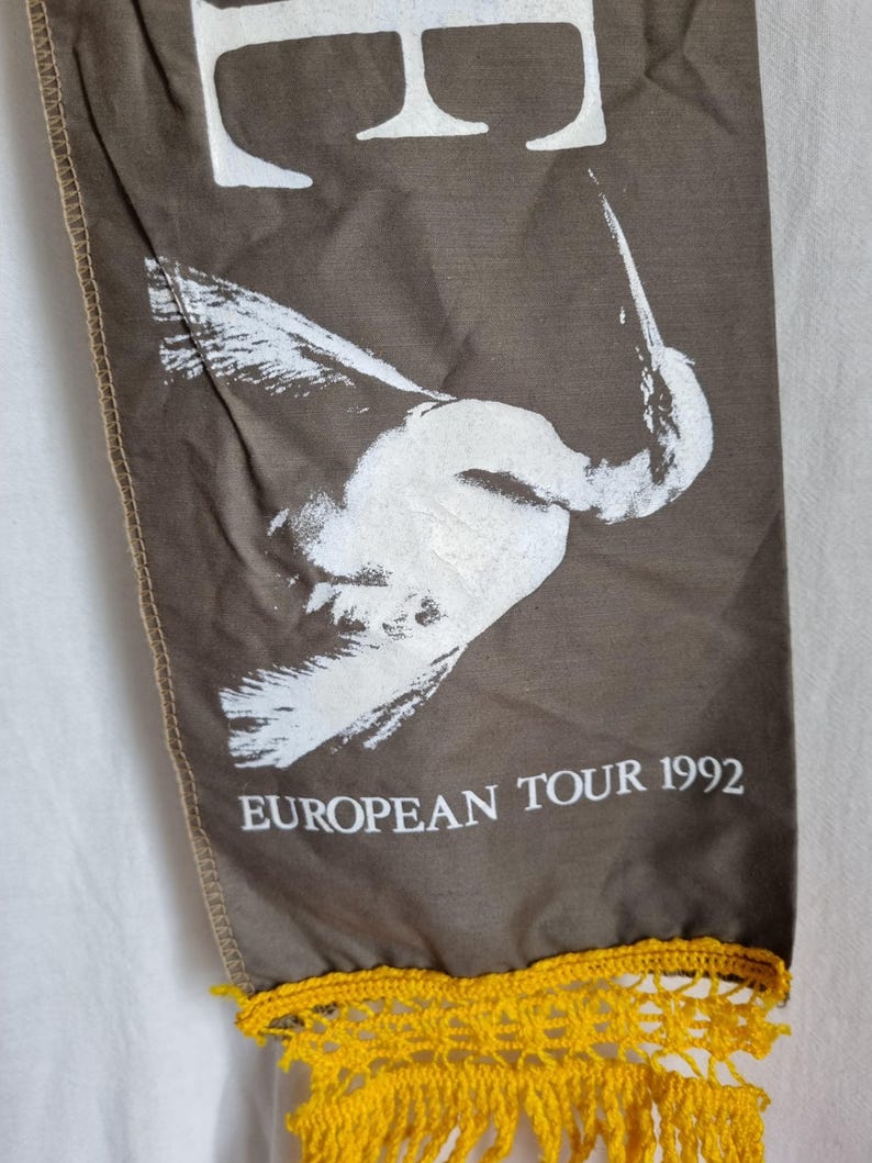 Puede incluir: Una pancarta de tela marr&oacute;n con un dise&ntilde;o de p&aacute;jaro blanco y el texto "EUROPEAN TOUR 1992". La pancarta tiene una franja de crochet amarilla.