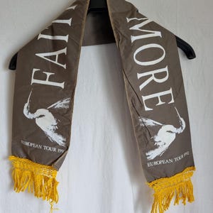 Puede incluir: Una bufanda de tela marr&oacute;n con texto blanco que dice "Faith No More" y "European Tour 1992". La bufanda tiene flecos amarillos en ambos extremos.