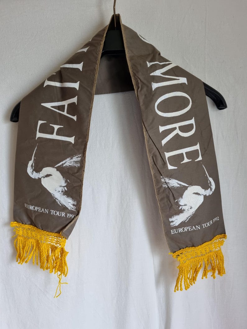 Puede incluir: Una bufanda de tela marr&oacute;n con texto blanco que dice "Faith No More" y "European Tour 1992". La bufanda tiene flecos amarillos en ambos extremos.