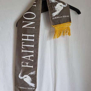 Puede incluir: Una bufanda de tela marr&oacute;n con texto blanco que dice "Faith No More" y un gr&aacute;fico de p&aacute;jaro blanco. La bufanda tiene flecos amarillos en ambos extremos y el texto "European Tour 1992" est&aacute; impreso en la esquina inferior derecha.