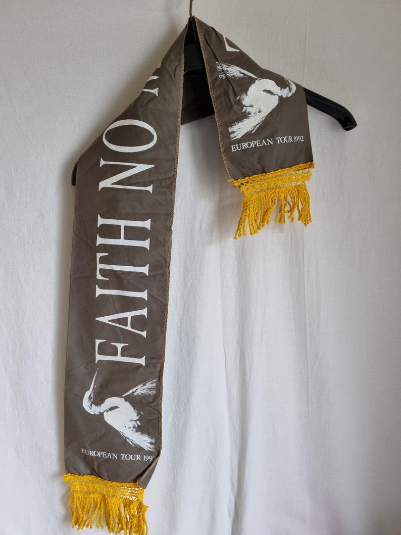 Puede incluir: Una bufanda de tela marr&oacute;n con texto blanco que dice "Faith No More" y un gr&aacute;fico de p&aacute;jaro blanco. La bufanda tiene flecos amarillos en ambos extremos y el texto "European Tour 1992" est&aacute; impreso en la esquina inferior derecha.