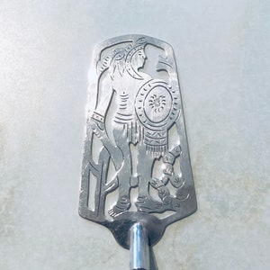 Puede incluir: Espátula de metal plateado con un diseño decorativo recortado de un guerrero sosteniendo un escudo. La espátula tiene un mango de madera oscura y una virola de metal plateado. El guerrero lleva un casco y una armadura.