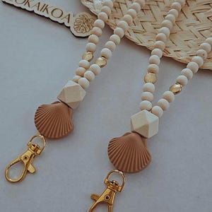 May include: Two beaded lanyards with wooden beads, geometric shapes, and seashell charms. Each lanyard has a gold-colored clasp. The lanyards are beige and brown. The text "TEOKAIKOA" is visible on a wooden tag.