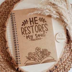 May include: A brown spiral bound journal with the words "HE RESTORES my soul" and "PSALM 23:3" engraved on the cover. The journal is decorated with a floral design and a book with flowers growing out of it. The journal is sitting on a woven brown surface.