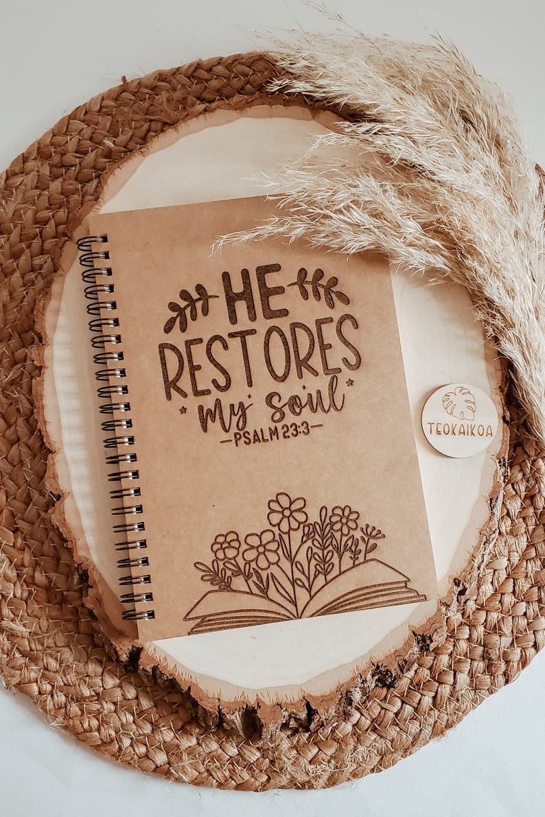 May include: A brown spiral bound journal with the words "HE RESTORES my soul" and "PSALM 23:3" engraved on the cover. The journal is decorated with a floral design and a book with flowers growing out of it. The journal is sitting on a woven brown surface.