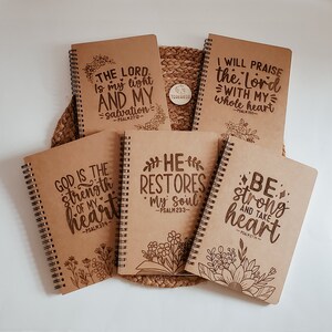 May include: Six brown spiral notebooks with inspirational quotes and floral designs. The quotes include "The Lord is my light and my salvation", "I will praise the Lord with my whole heart", "God is the strength of my heart", "He restores my soul", and "Be strong and take heart".