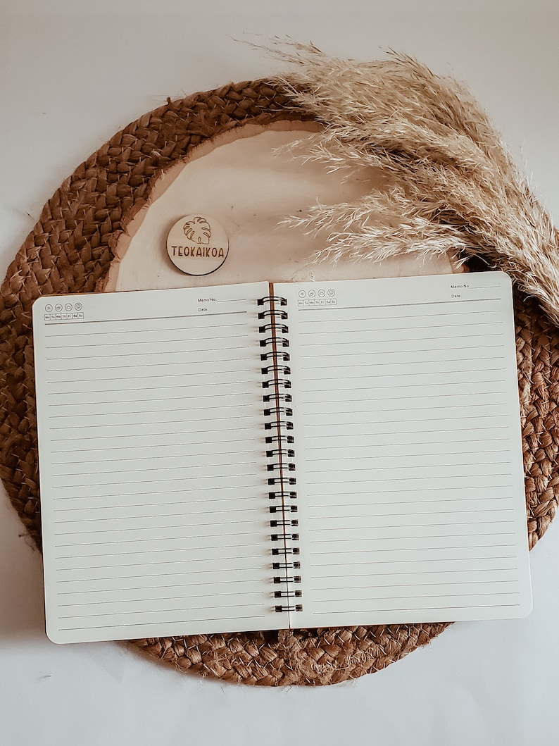 May include: An open spiral bound notebook with lined pages, resting on a woven straw mat. A wooden round tag with the text "TEOKAIKOA" is visible on the mat.