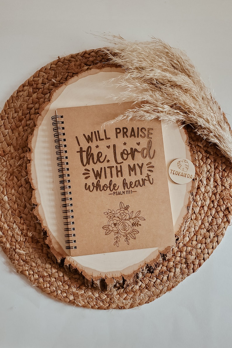May include: A brown notebook with a spiral binding and a quote that reads "I will praise the Lord with my whole heart - Psalm 111:1" with a floral design at the bottom. The notebook is sitting on a woven mat with a small wooden tag that reads "TEOKAI KOA".