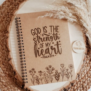 May include: A brown spiral notebook with the text "God is the strength of my heart - Psalm 37:4" and a floral design at the bottom. The notebook is on a woven surface with a wooden tag that says "Teoakikoa".