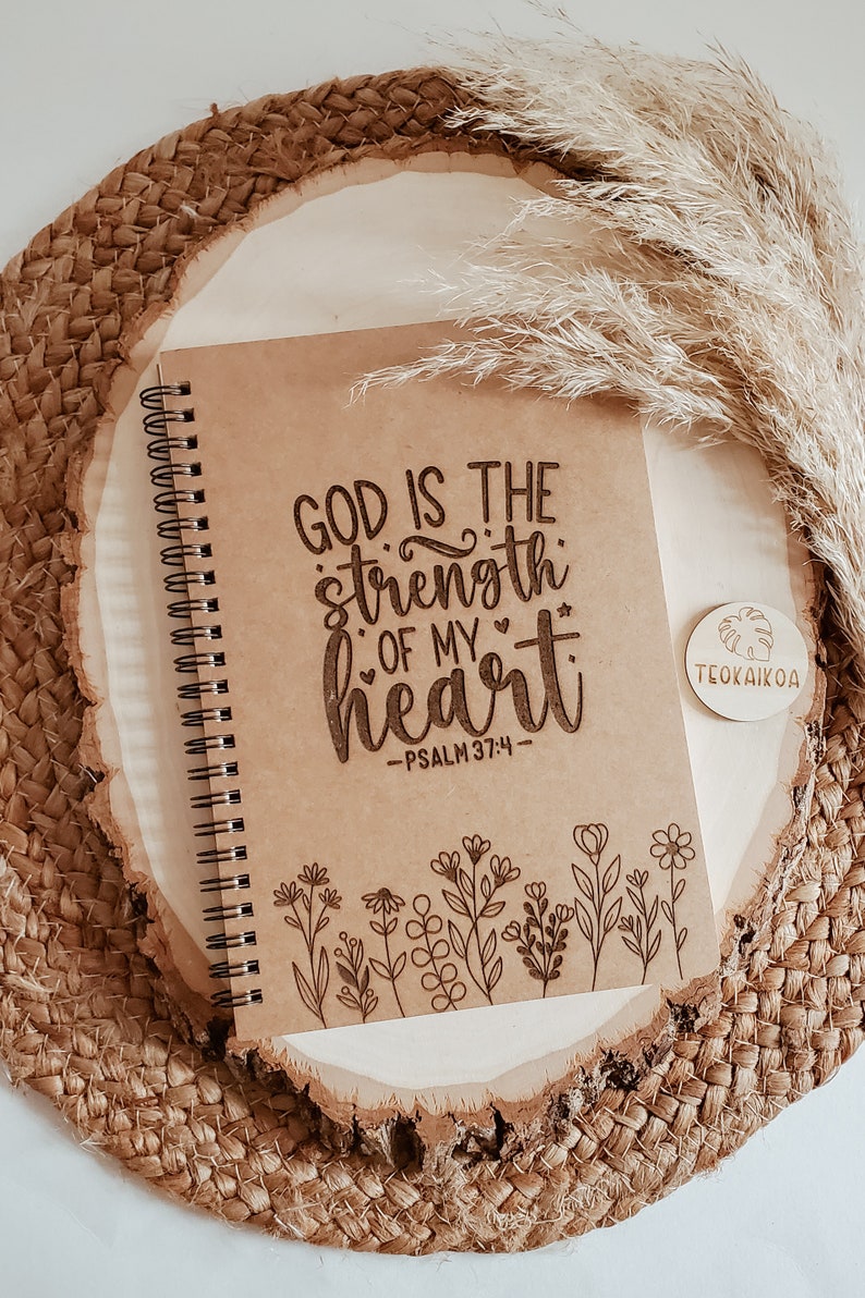 May include: A brown spiral notebook with the text "God is the strength of my heart - Psalm 37:4" and a floral design at the bottom. The notebook is on a woven surface with a wooden tag that says "Teoakikoa".