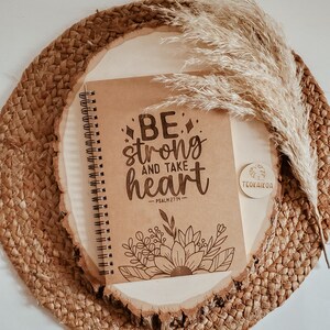 May include: A brown notebook with a spiral binding and the words "BE strong and take heart - PSALM 27:14" printed on the cover. The notebook is on a wooden surface with a brown woven mat and a dried grass plume.
