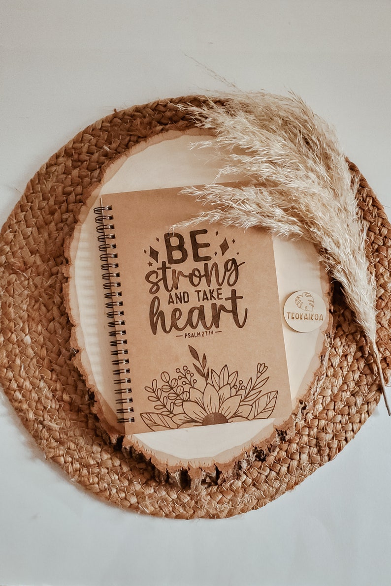 May include: A brown notebook with a spiral binding and the words "BE strong and take heart - PSALM 27:14" printed on the cover. The notebook is on a wooden surface with a brown woven mat and a dried grass plume.