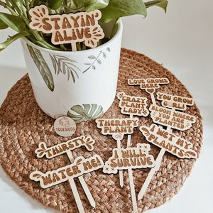 May include: A set of wooden plant markers with various phrases, including "Stayin' Alive", "Love Grows Here", "Crazy Plant Lady", "Therapy Plant", "You Grow Girl", "Bloom Where You Are Planted", "I Wet My Plants", "Thirsty", "Water Me", and "I Will Survive".