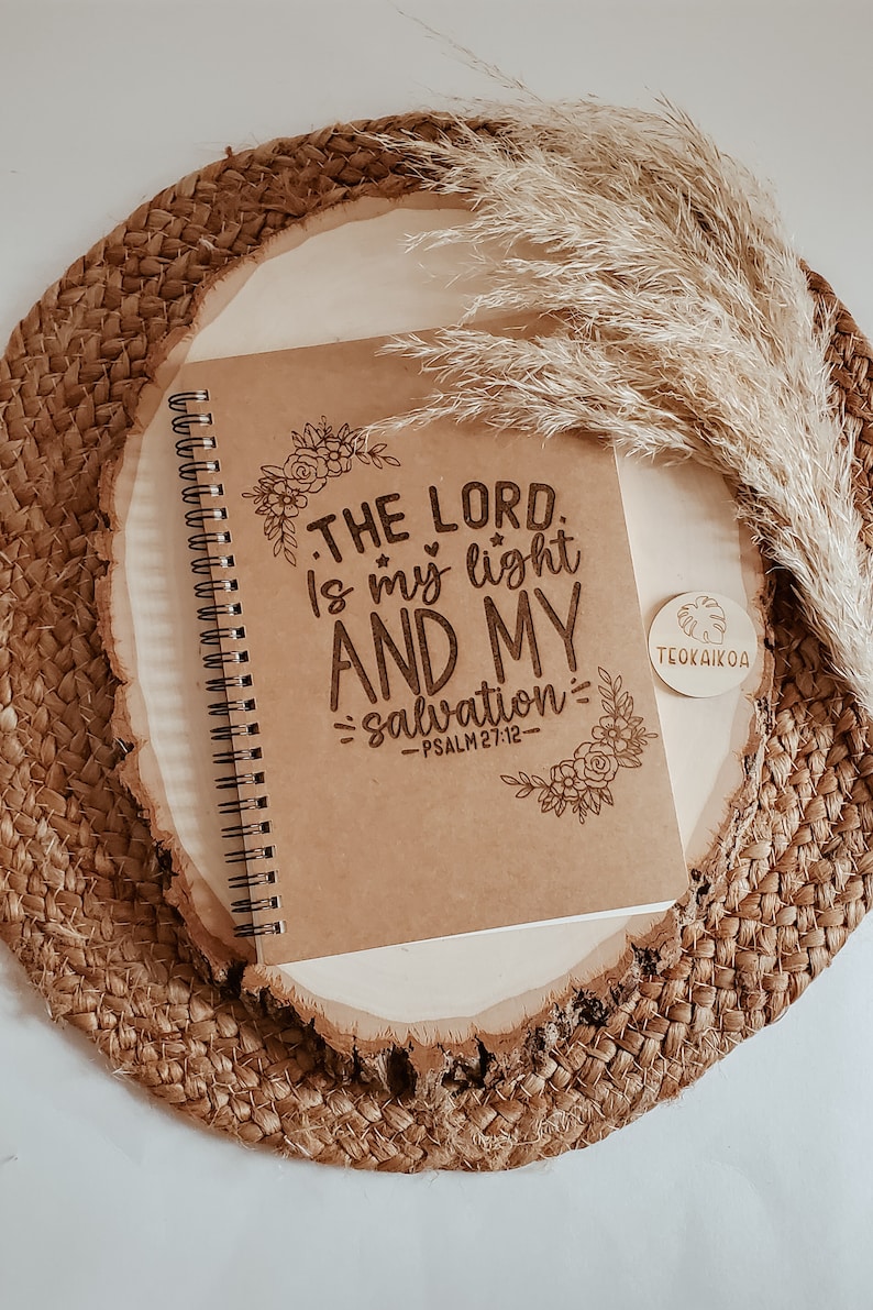 May include: A brown spiral notebook with the text "THE LORD is my light AND MY salvation - PSALM 27:12" in a decorative font. The notebook is on a woven round surface with a small wooden tag that says "TEOKAIKOA".