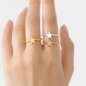 Peut inclure: Trois bagues en forme d'étoile, dorées, argentées et or rose. Chaque bague présente un motif d'étoile et un anneau perlé. Les bagues sont présentées sur une main, mettant en valeur les différentes couleurs et styles.