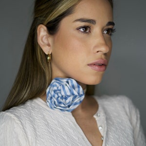 May include: A woman with long brown hair wearing a white knitted cardigan with blue and white striped fabric flower on her neck.