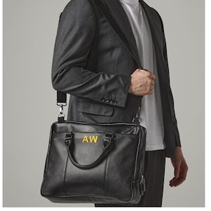 May include: Black leather briefcase with a shoulder strap. The briefcase has a top handle and a front pocket. The initials "AW" are in gold lettering on the front of the bag. The person is wearing a gray suit and a white shirt.