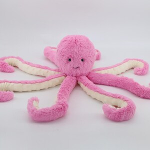 Puede incluir: Un peluche de pulpo rosa con ocho largos tentáculos curvos. Los tentáculos son rosas por fuera y color crema por dentro. El pulpo tiene una cabeza redonda con pequeños ojos bordados y una sonrisa cosida.