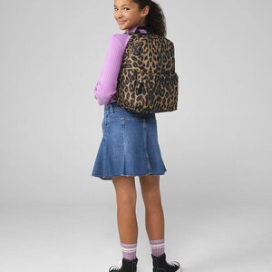 May include: A leopard print backpack with black accents. The backpack is worn by a person wearing a light purple long-sleeve shirt, a denim skirt, black high-top sneakers, and striped socks. The background is a solid light gray.