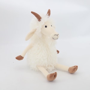 Puede incluir: Un peluche de cabra con cuerpo color crema y pelaje lanudo. Tiene cuernos, pezuñas y una cara sonriente marrones. El juguete está sentado con las piernas extendidas, creando una apariencia juguetona y tierna.