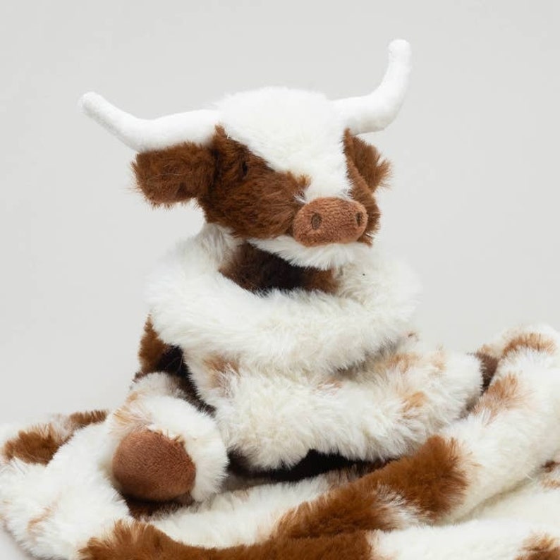 May include: A plush stuffed animal shaped like a highland cow with soft brown and white fur. The toy features two white horns, a brown nose, and a long, flowing white and brown patterned tail or blanket-like attachment extending from its body.