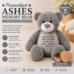 Könnte beinhalten: Ein grauer Plüschbär mit einem glatten, hellgrauen Bauchfleck, bestickt mit dem Text: Hold me close when you need me the most, Always with you, Nanny, x. Neben dem Bären steht eine offene, herzförmige Erinnerungsdose aus roségoldfarbenem Metall.