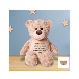 Puede incluir: Un oso de peluche beige con un mensaje bordado en su pecho que dice "Hold me close when you need me the most Always with you Nanny x". El oso está sentado sobre un fondo azul con estrellas blancas. Una pequeña caja de corazón de oro rosa se encuentra en la esquina inferior derecha de la imagen.