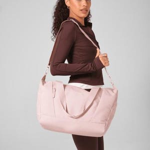 Può includere: Una borsa da viaggio rosa chiaro con tracolla e tasca con cerniera. La borsa è portata da una persona che indossa un top sportivo marrone e leggings. Lo sfondo è grigio chiaro.