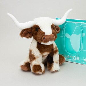 May include: A plush longhorn cow toy with brown and white fur, featuring long white horns and dark eyes. Beside it sits a teal gift bag with the text "amanda" and a small sheep graphic printed on the front, featuring teal rope handles.