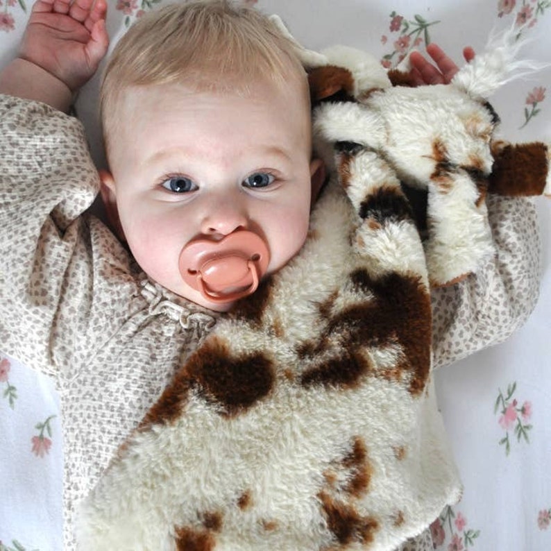 May include: A baby with light hair and blue eyes wearing a beige long-sleeved shirt with a small brown pattern. The baby has a solid peach-colored pacifier in their mouth and is holding a plush security blanket with a white and brown cow print.