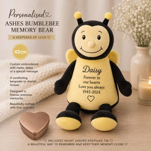 Könnte beinhalten: Ein Plüsch-Hummel-Gedenkbär aus gelbem und schwarzem Stoff mit dem gestickten Namen Daisy, dem Text Forever in our hearts Love you always 1945-2024 und einem Herzsymbol. Eine kleine herzförmige Erinnerungsdose aus Metall steht daneben.