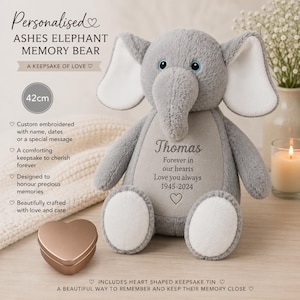 Könnte beinhalten: Ein grauer Plüschelefant mit großen Ohren und weißen Akzenten. Auf dem Bauch befindet sich eine Stickerei: Thomas, Forever in our hearts, Love you always, 1945-2024, sowie ein Herzsymbol. Daneben steht eine kleine herzförmige Metalldose als Andenken.