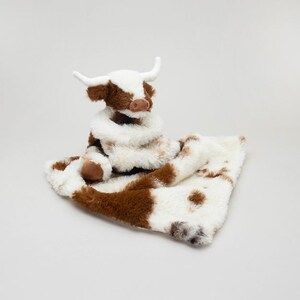 May include: A plush stuffed animal shaped like a highland cow with white and brown fur, featuring two white horns and a brown nose. The cow is attached to a soft, fuzzy blanket with a white and brown cow-print pattern made of plush material.