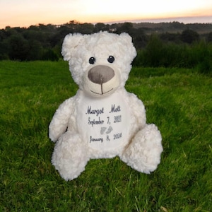 Könnte beinhalten: Weißer Plüschteddybär mit gesticktem Text: "Margot Mott, 7. September 2021, 8. Januar 2024" und einem Paar Fußabdrücken.
