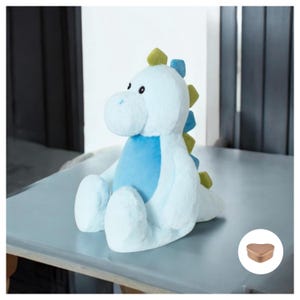 May include: A light blue plush dinosaur toy with green accents. The dinosaur is sitting on a table with a small heart-shaped box in the corner.